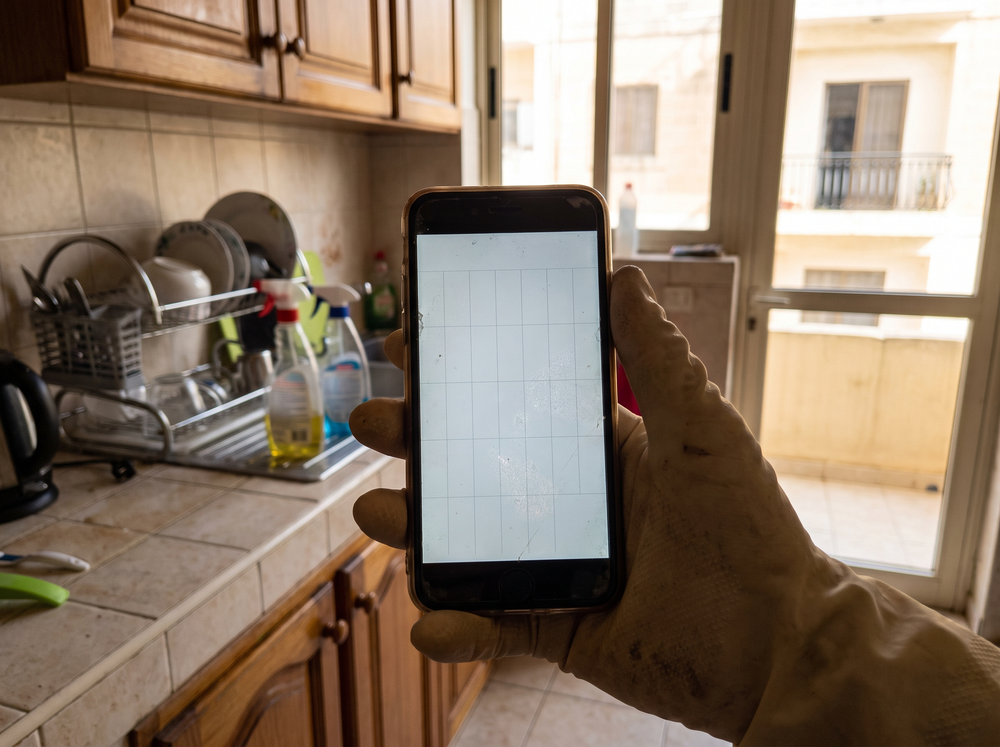 Cleaner's hand holding a phone showing a blank calendar app, Maltese apartment kitchen in the background